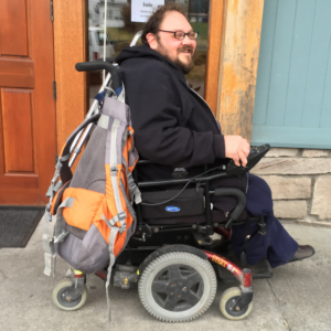 Man with a beard and black sweatshirt uses a wheelchair on the sidewalk in front of a building